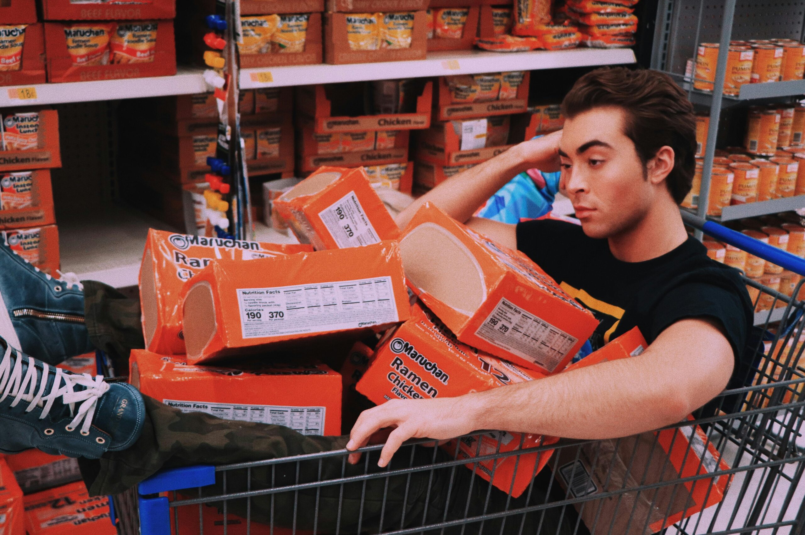 A man lounging in a shopping cart filled with Maruchan ramen in a supermarket aisle.