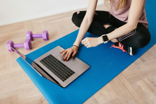 Crop female in sportswear sitting on blue yoga mat on floor and surfing internet on laptop