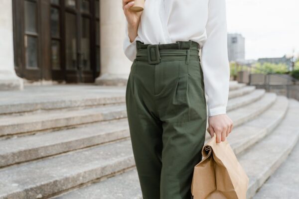 Woman in stylish outfit holding a takeaway coffee and paper bag on outdoor steps.